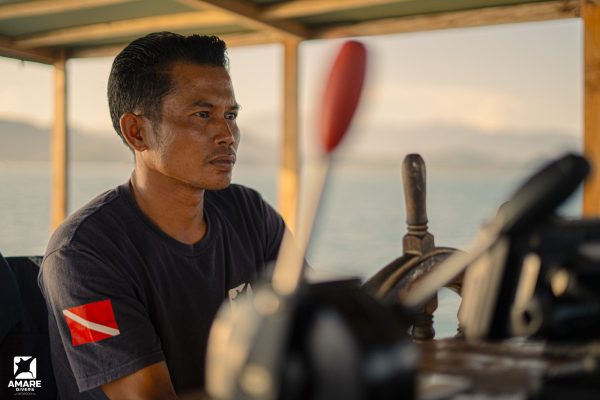 A profile portrait of a weathered Indonesian boat captain wearing a navy blue Amare Divers uniform, smiling warmly while steering the Maria Seascape liveaboard in Komodo National Park.
