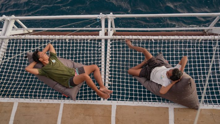 A view from the dive deck of the Maria Seascape liveaboard, looking out over the blue water and iconic karst islands of Komodo National Park, with scuba diving equipment neatly arranged on racks.